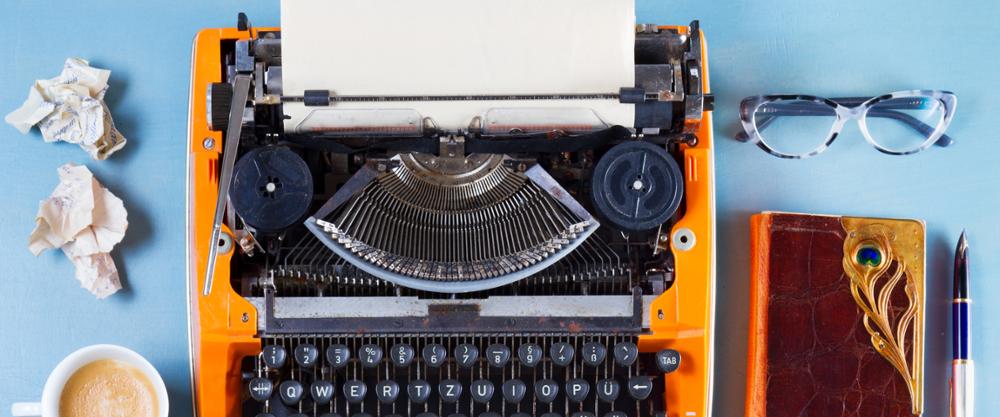 Máquina de escribir sobre una mesa, al lado hay un café, unas gafas, un cuaderno y una pluma