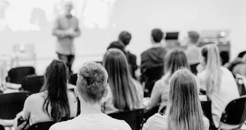 Fotografía difuminada en blanco y negro tomada por detrás en la que se ve unos alumnos sentados y un profesor dando clase 