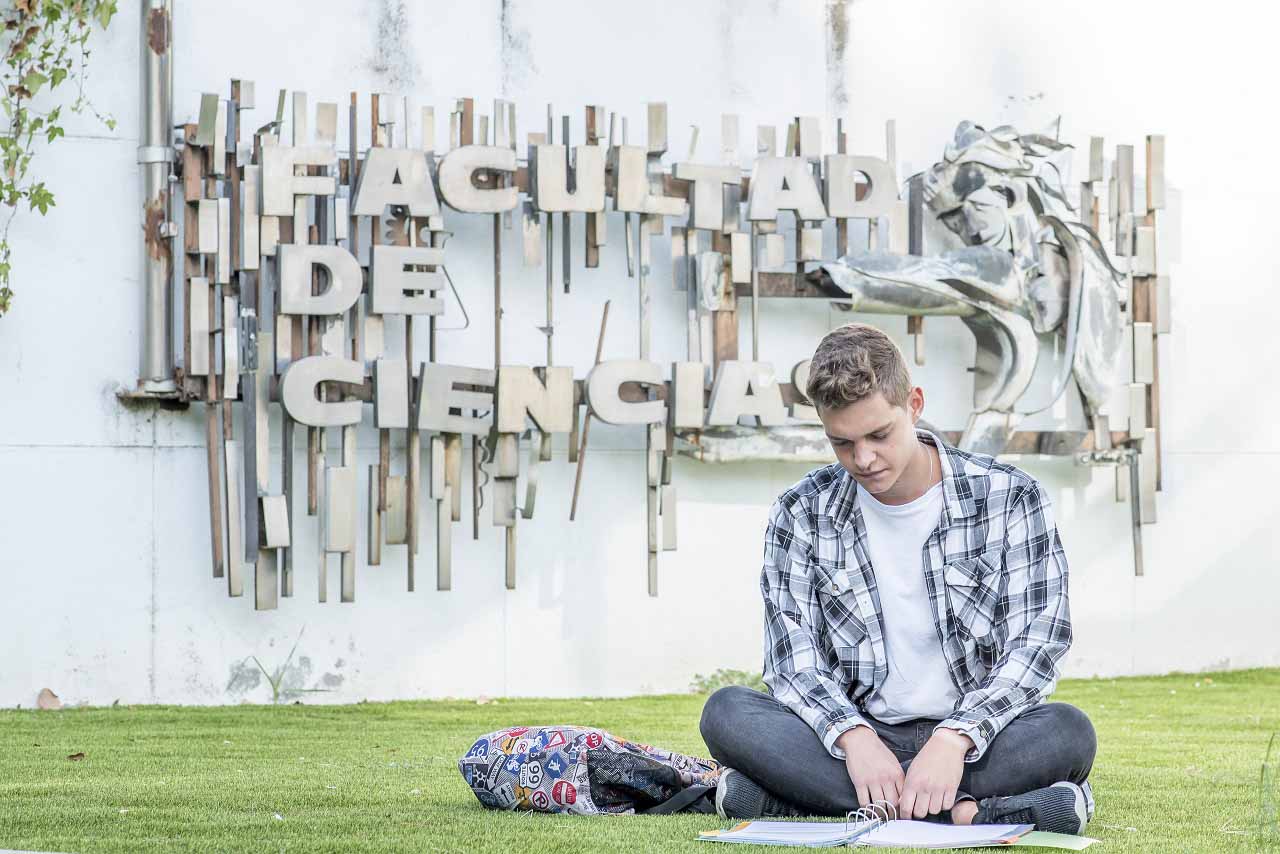 Un estudiante lee sentado en el césped, delante de las letras que indican la Facultad de Ciencias