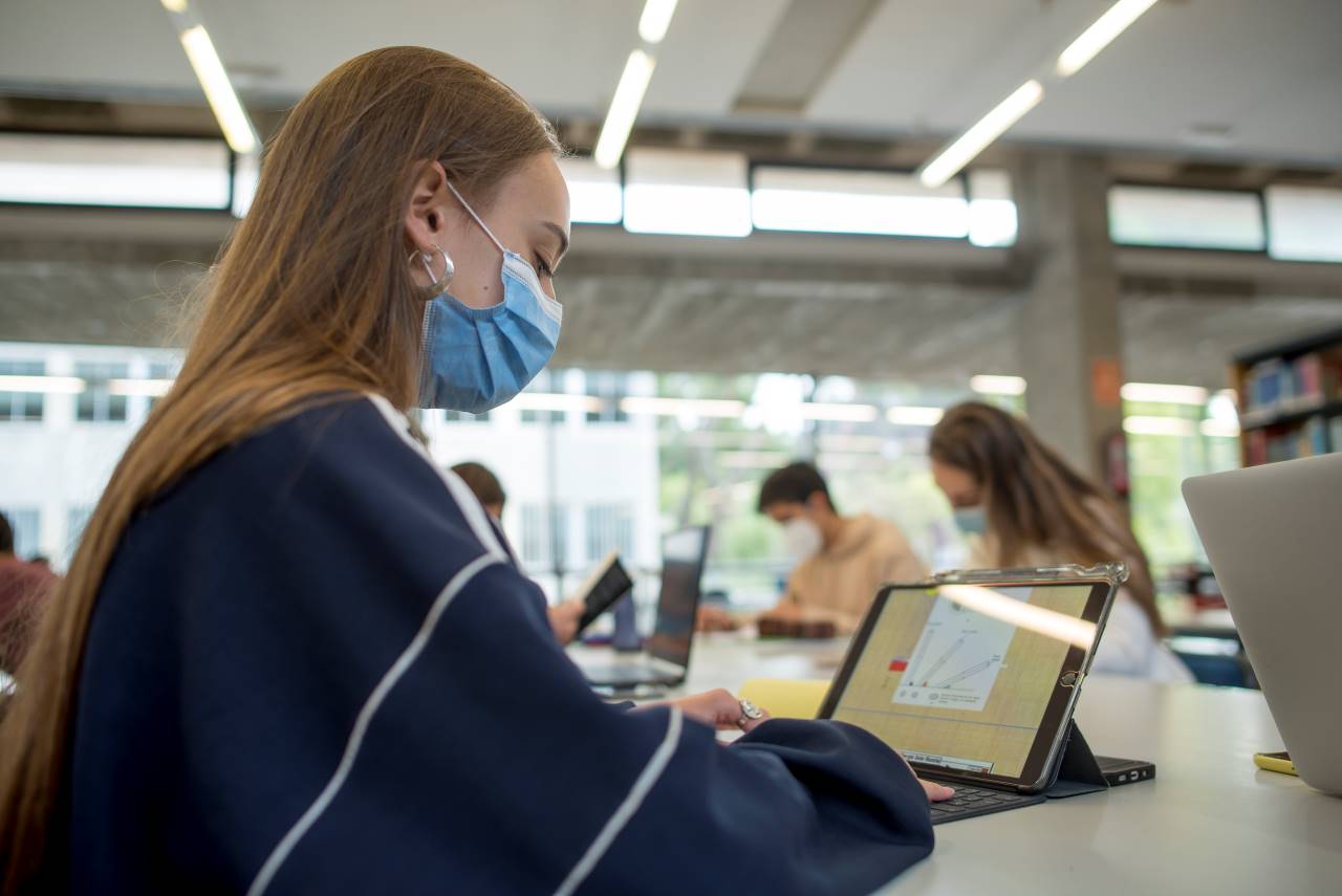Imagen que muestra a una estudiante con mascarilla leyendo en un ordenador portátil en la biblioteca