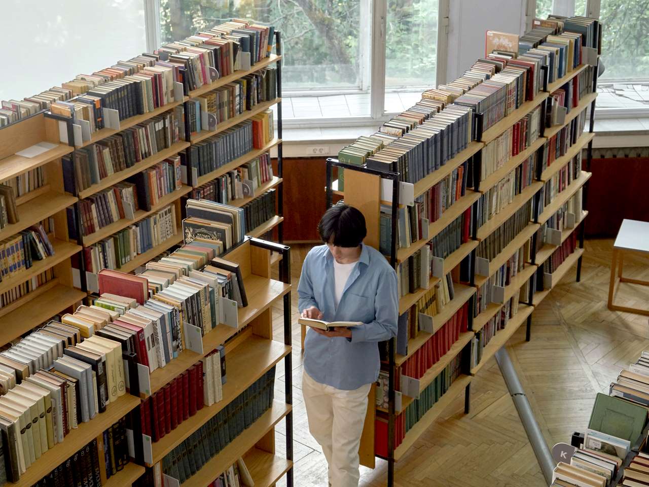 Un estudiante lee un libro de pie apoyado en una estantería de una biblioteca.
