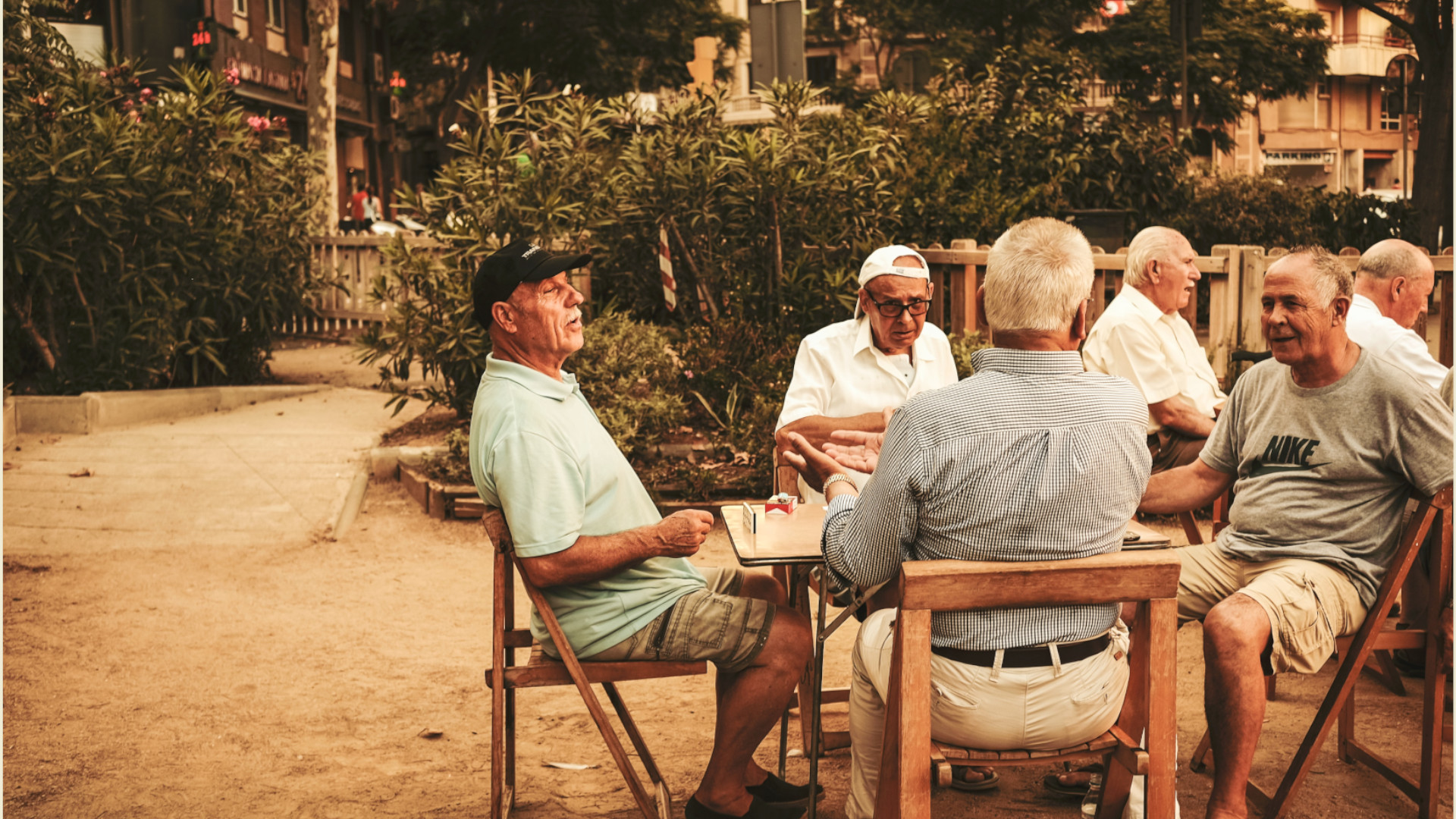 Grupo de tres hombres mayores sentadas al aire libre en torno a una mesa de ajedrez con ropa de verano y actitud relajada. 
