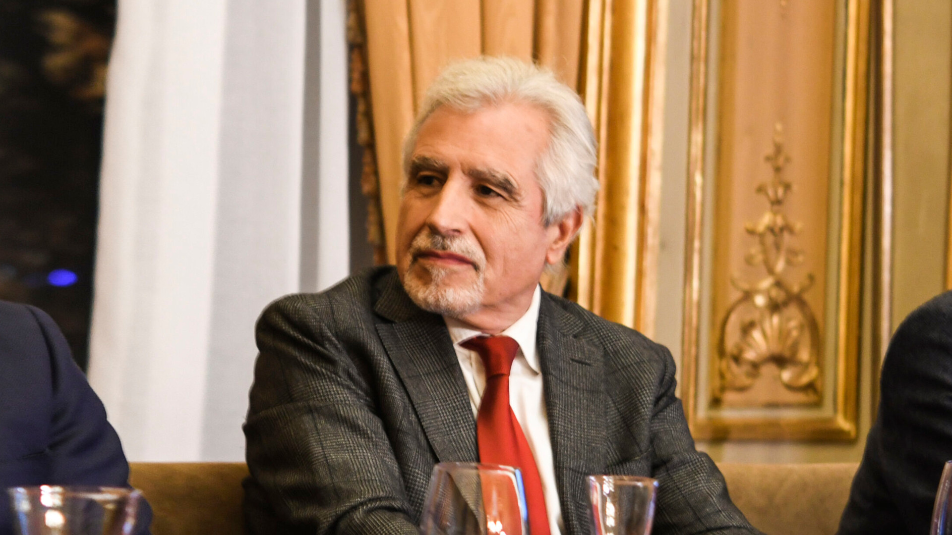 Fotografía de medio cuerpo de un hombre de cabello canoso con traje oscuro y corbata roja detrás de una mesa de conferencias con fondo de cortinas blancas y pared dorada.