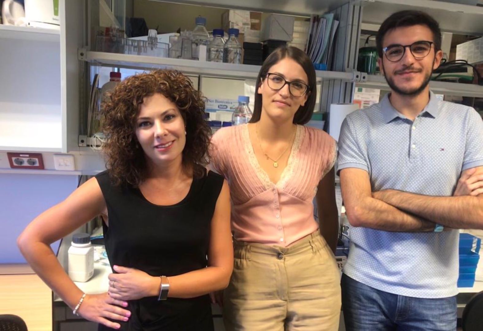 Fotografía de tres cuartos de la entrevistada con dos alumnos, una chica y un chico a su izquierda apoyados en una mesa de laboratorio.