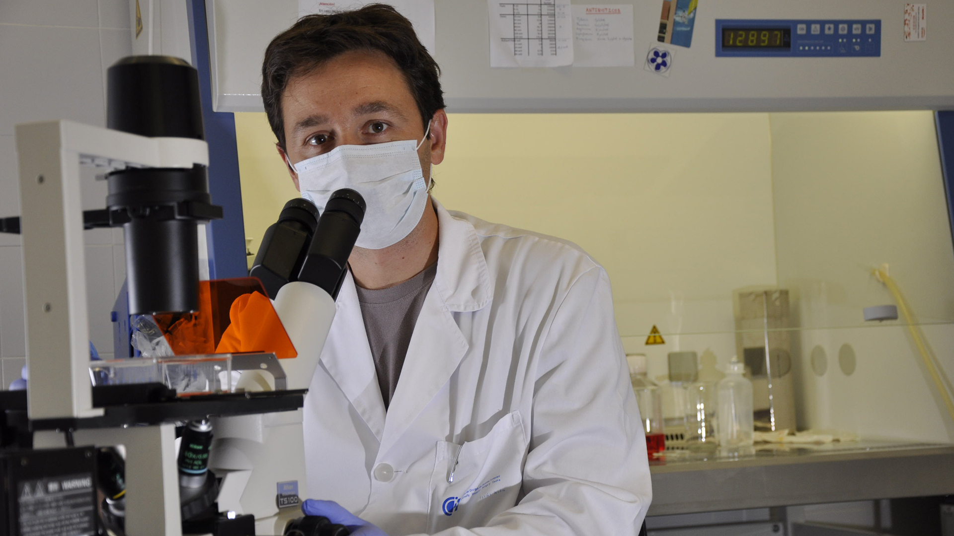 Fotografía de un hombre de pelo corto con bata blanca levantando la vista de un microscopio.