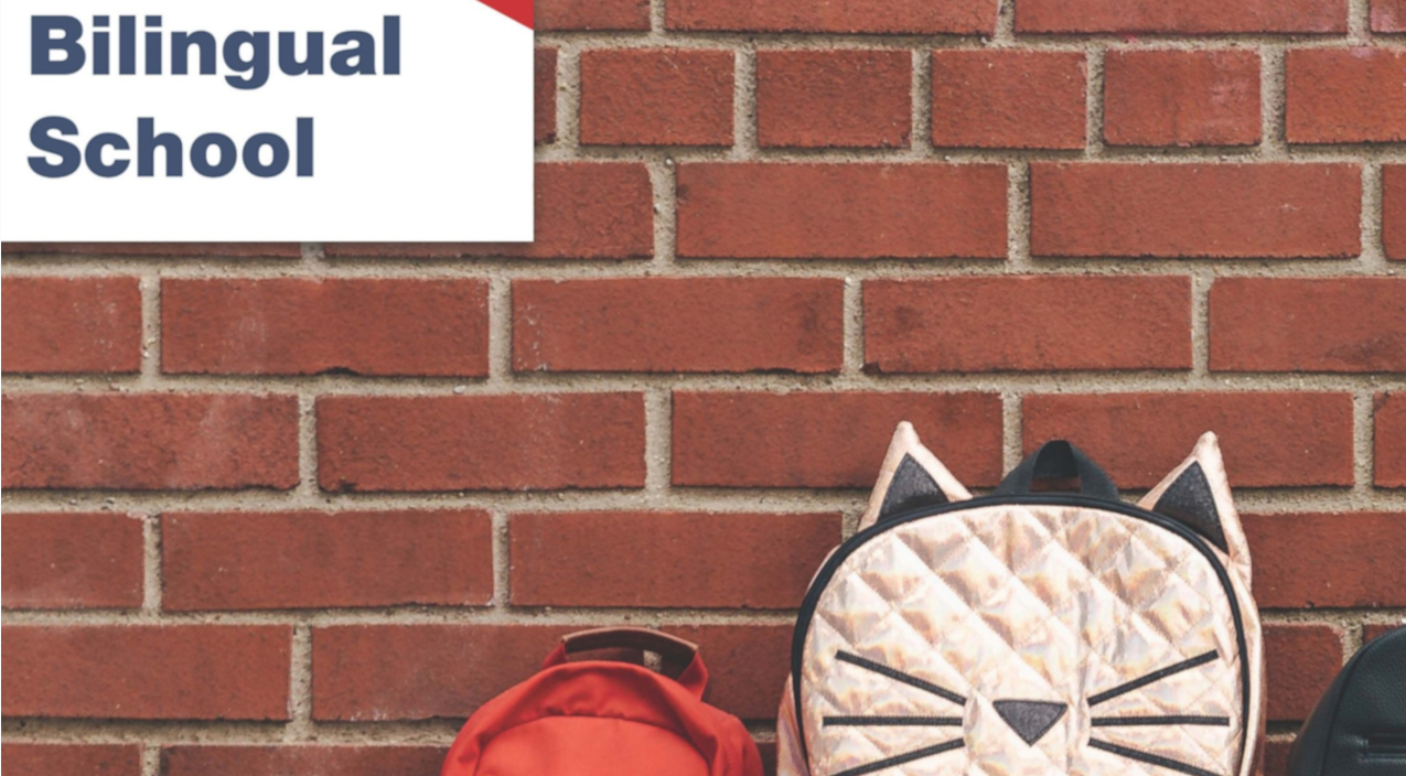 Tres mochilas infantiles apoyadas en el suelo sujetas sobre una pared de ladrillo rojo con un cartel de una escuela bilingüe.