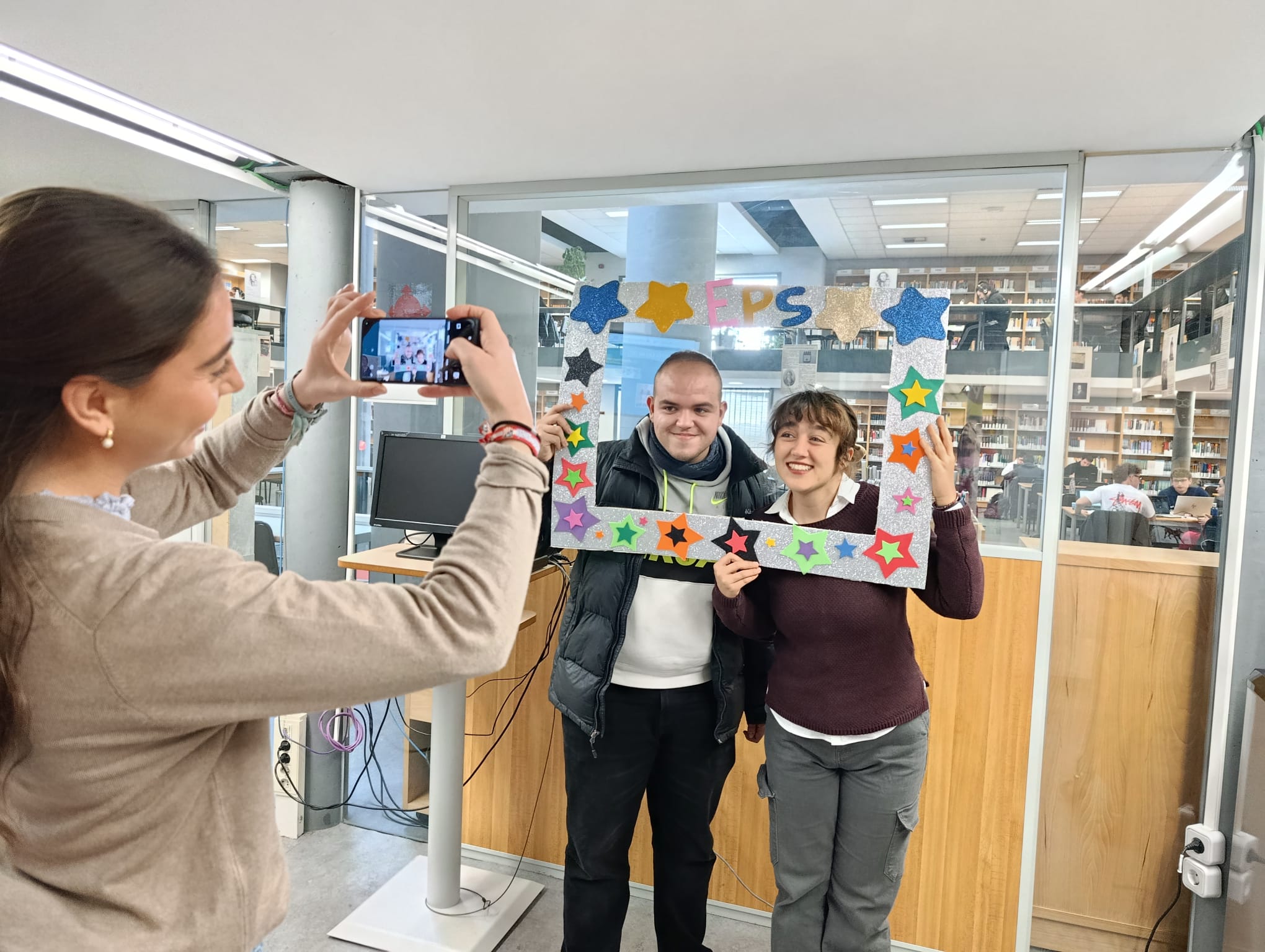 Imagen de una alumna haciendo una foto a dos alumnos con un marco de la Biblioteca EPS