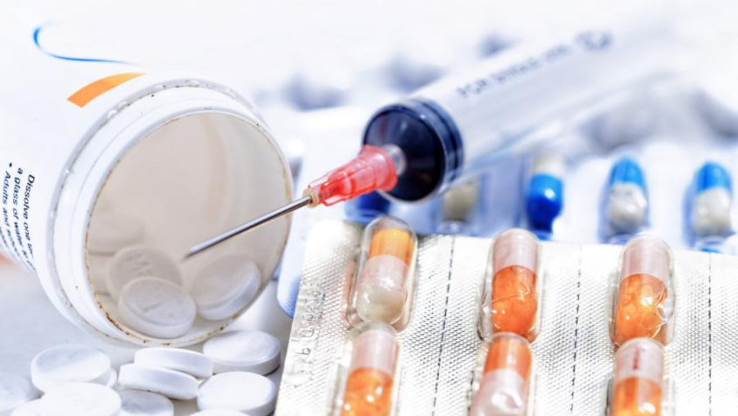 Medicamentos variados: pastillas blancas, cápsulas en blíster y una jeringa sobre superficie clínica